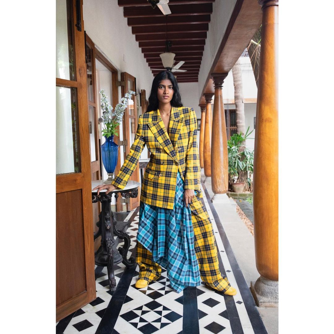 Woman in yellow plaid blazer and blue plaid skirt poses indoors on a black and white geometric floor near dark wood doors.