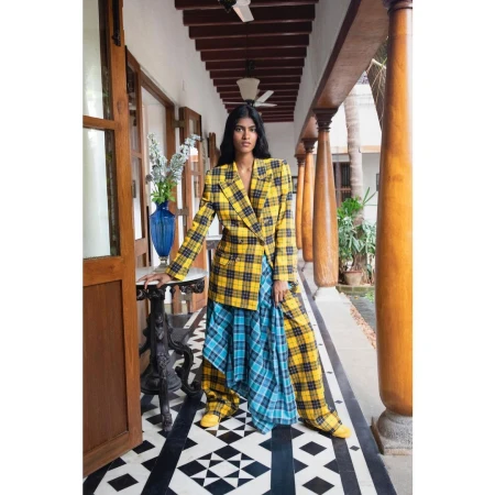 Woman in yellow plaid blazer and blue plaid skirt poses indoors on a black and white geometric floor near dark wood doors.