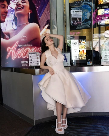 Adult woman in a white dress poses indoors against a movie poster and concession stand background, smiling and holding her head.