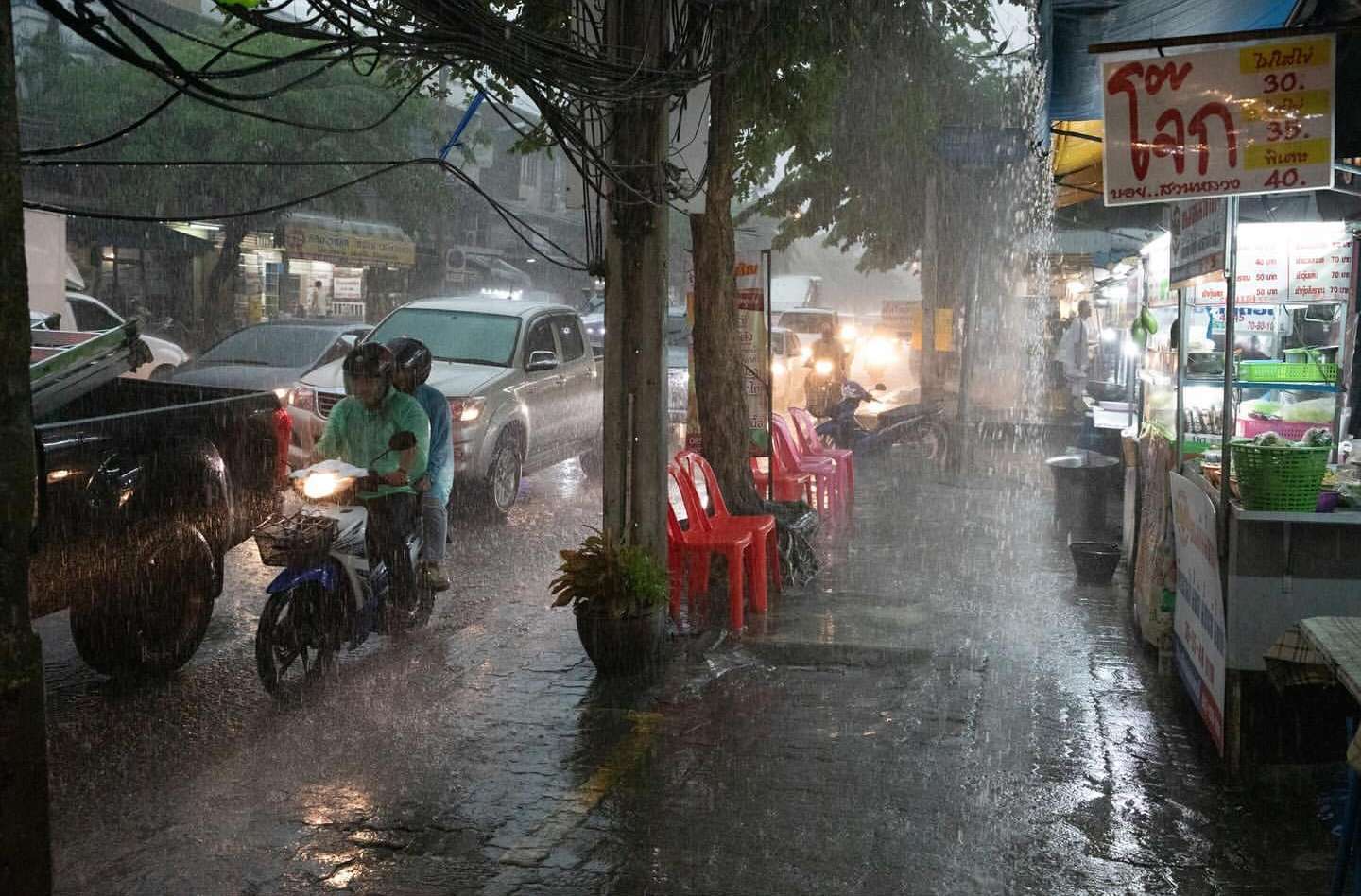 Heavy rain showers on a street scene with people riding a scooter, parked cars, red plastic chairs, and a food stand along a sidewalk.