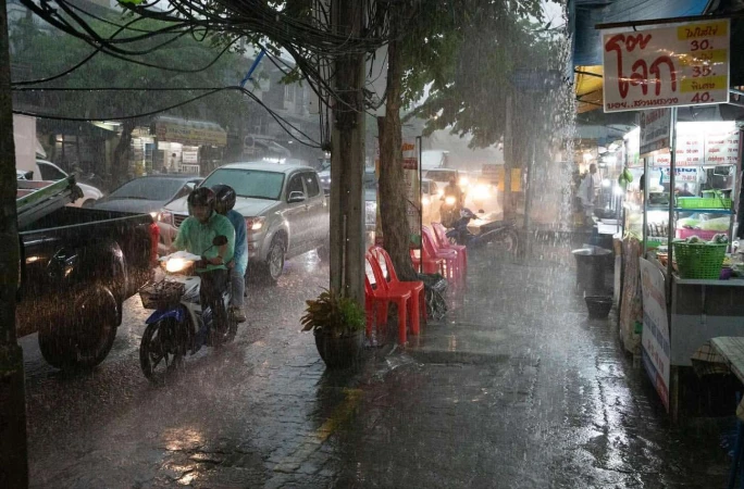 Heavy rain showers on a street scene with people riding a scooter, parked cars, red plastic chairs, and a food stand along a sidewalk.