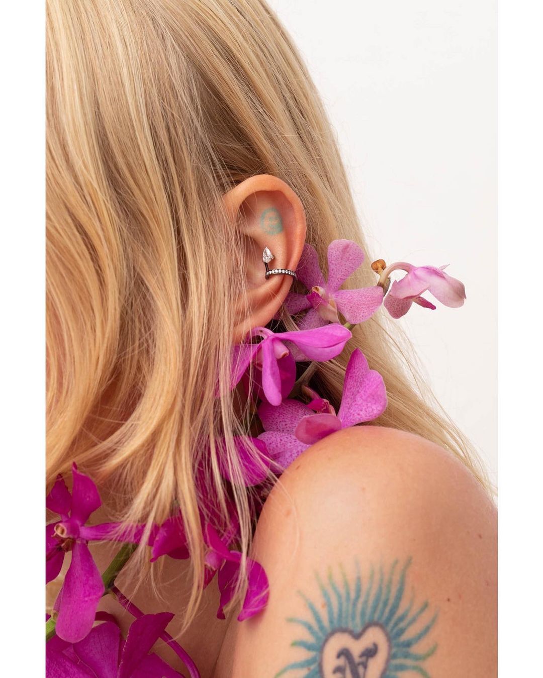 Close-up of a blonde woman's ear adorned with a teardrop helix piercing and draped with vibrant pink orchids, set against a white backdrop.