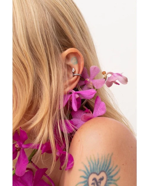 Close-up of a blonde woman's ear adorned with a teardrop helix piercing and draped with vibrant pink orchids, set against a white backdrop.