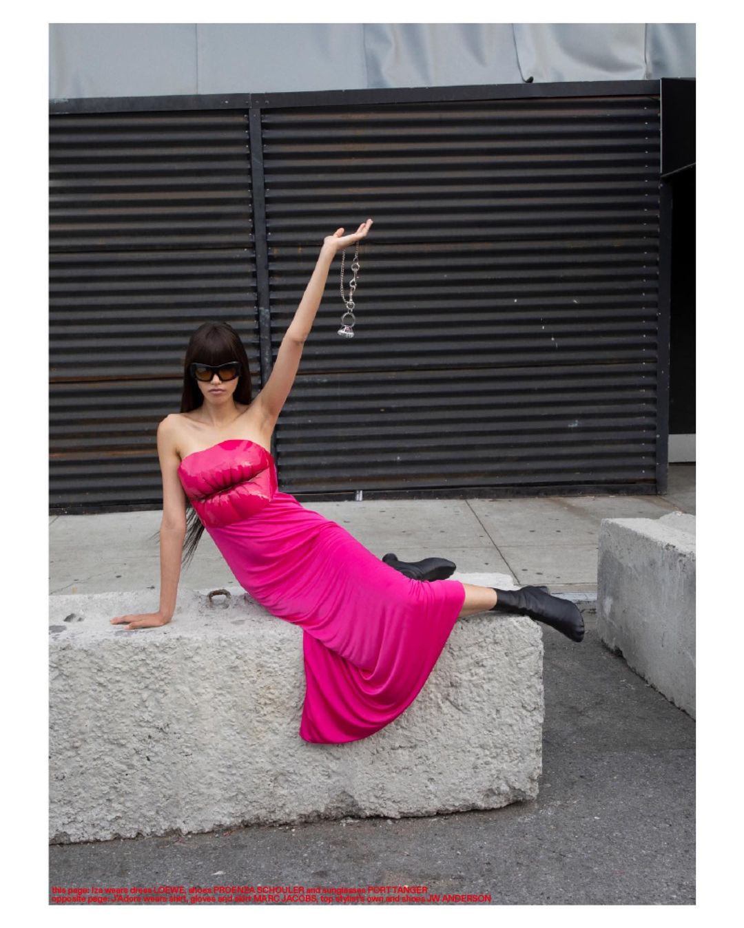 A woman with dark hair poses outdoors wearing a pink dress, black boots, and sunglasses while holding a chain with charms.