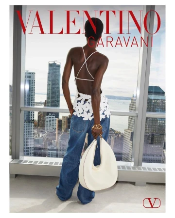 Woman in denim pants and a floral top holding a white handbag by a window overlooking a city skyline on an overcast day.