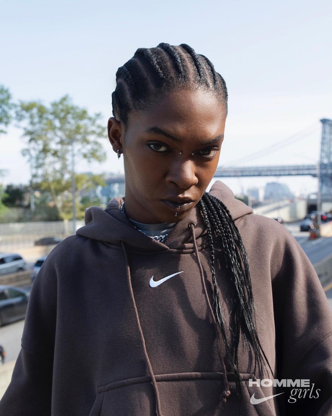 Woman in a brown hoodie with braided hair, an urban backdrop featuring a bridge and city buildings is visible in the background.