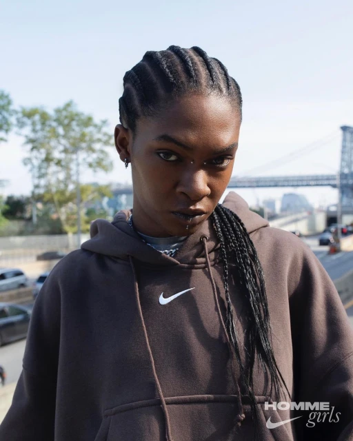 Woman in a brown hoodie with braided hair, an urban backdrop featuring a bridge and city buildings is visible in the background.