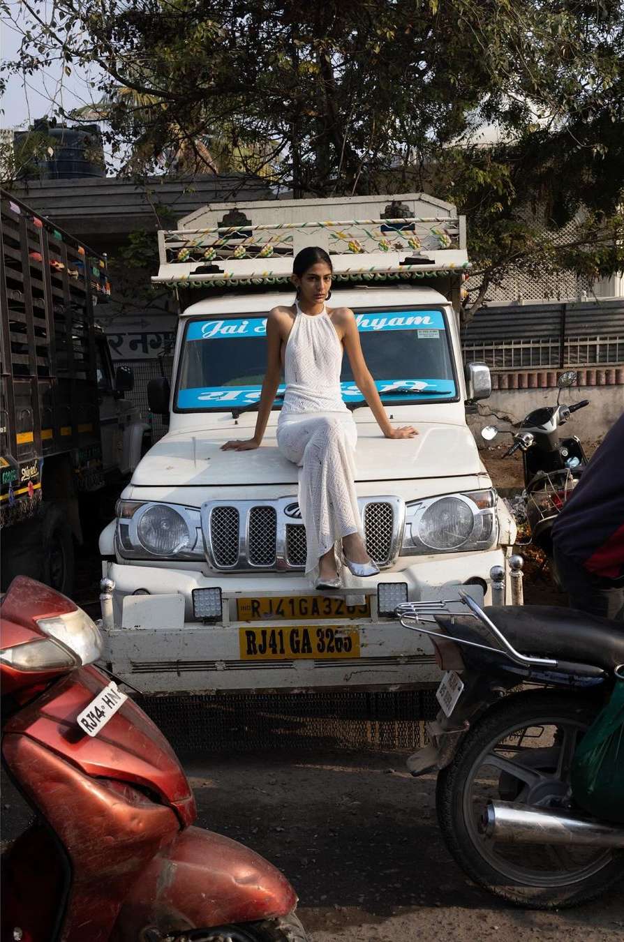 Young woman in white outfit poses on a white truck in an urban street setting with motorcycles and parked vehicles.