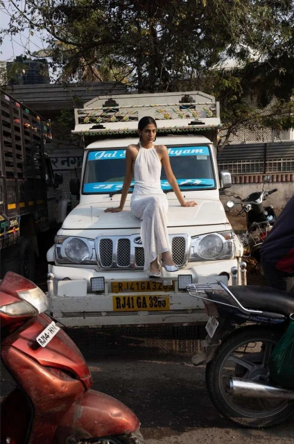 Young woman in white outfit poses on a white truck in an urban street setting with motorcycles and parked vehicles.