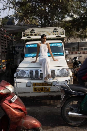 Young woman in white outfit poses on a white truck in an urban street setting with motorcycles and parked vehicles.