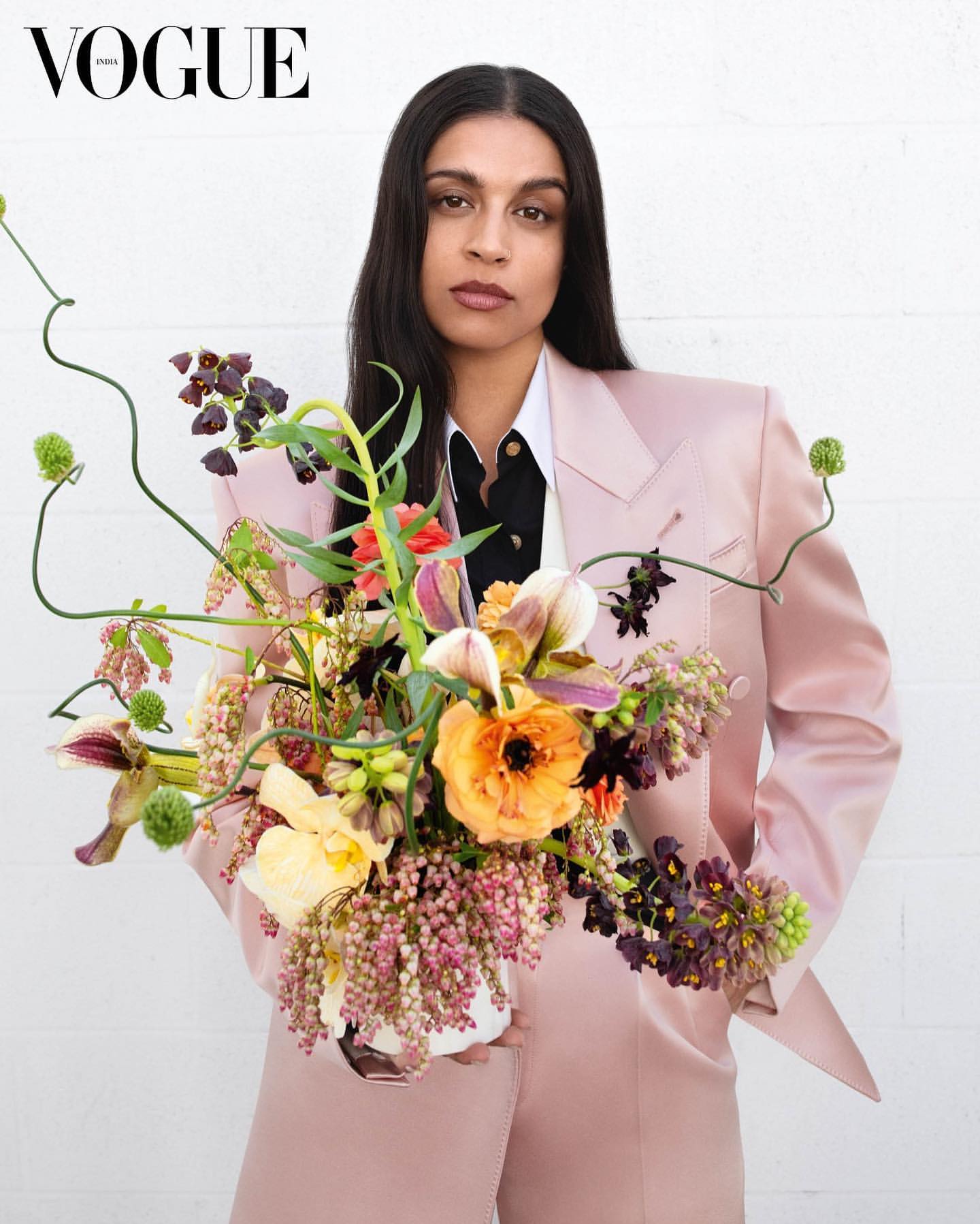 Portrait of a brunette adult woman in a pink blazer, holding a colorful bouquet in front of a white wall. Vogue logo visible.