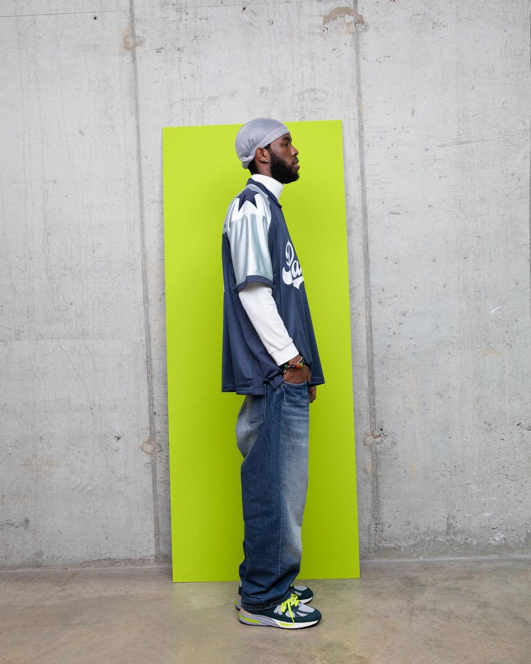Man in a sports jersey and baggy jeans standing in front of a lime green panel against a concrete wall in a medium shot.
