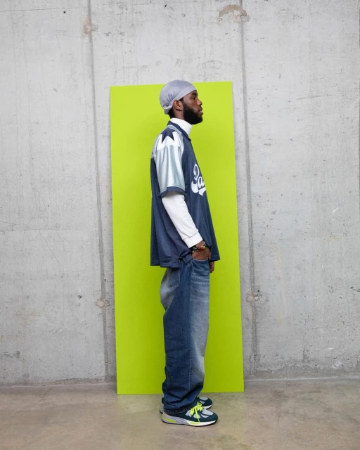 Man in a sports jersey and baggy jeans standing in front of a lime green panel against a concrete wall in a medium shot.