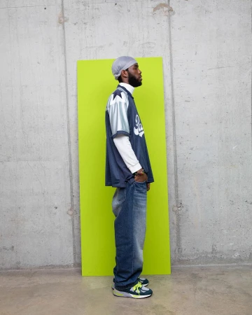 Man in a sports jersey and baggy jeans standing in front of a lime green panel against a concrete wall in a medium shot.