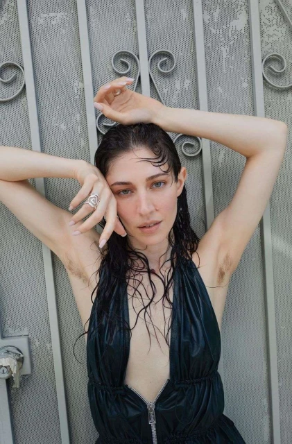 Woman poses against metal gate with hands raised, wet dark hair, wearing black dress. Outdoor fashion portrait. Ring is visible.