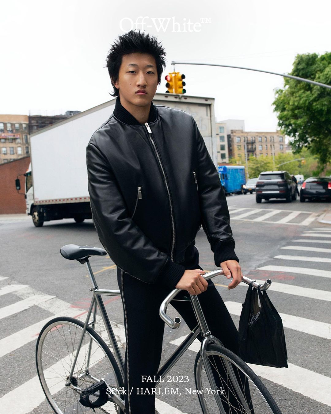 Asian man in a black jacket and pants, standing with a bicycle on a city street in New York, near a white truck and traffic lights.