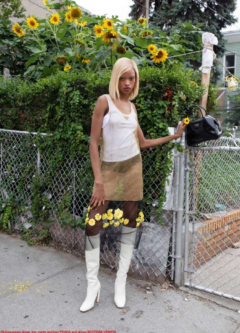 Woman poses in a sleeveless top, skirt, tights with yellow flowers, and white boots, holding a yellow flower and a black purse.