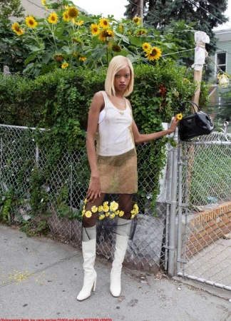 Woman poses in a sleeveless top, skirt, tights with yellow flowers, and white boots, holding a yellow flower and a black purse.