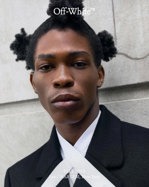 Close-up portrait of a Black male model with unique hairstyle wearing a black coat and white tie, posing in front of a neutral-toned wall.