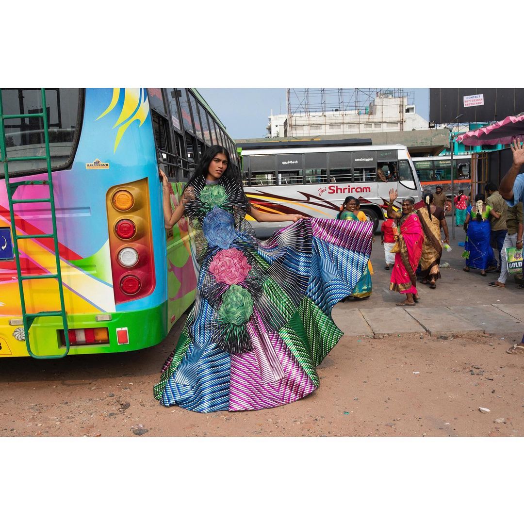 A woman in a long, colorful dress stands next to a bus at an outdoor bus stop, surrounded by other buses and pedestrians in daylight.