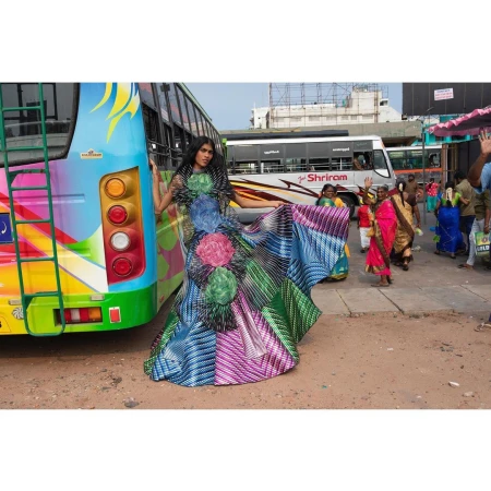 A woman in a long, colorful dress stands next to a bus at an outdoor bus stop, surrounded by other buses and pedestrians in daylight.