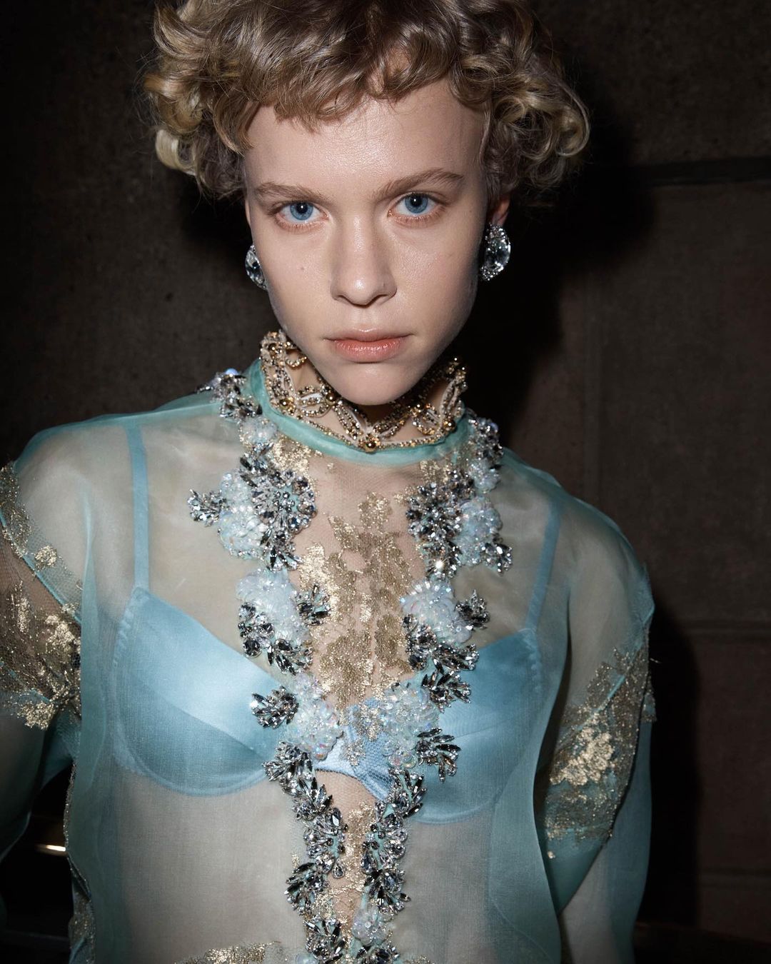 Close-up portrait of a young woman with blonde curly hair wearing a sheer aqua top with jewel embellishments and statement earrings.