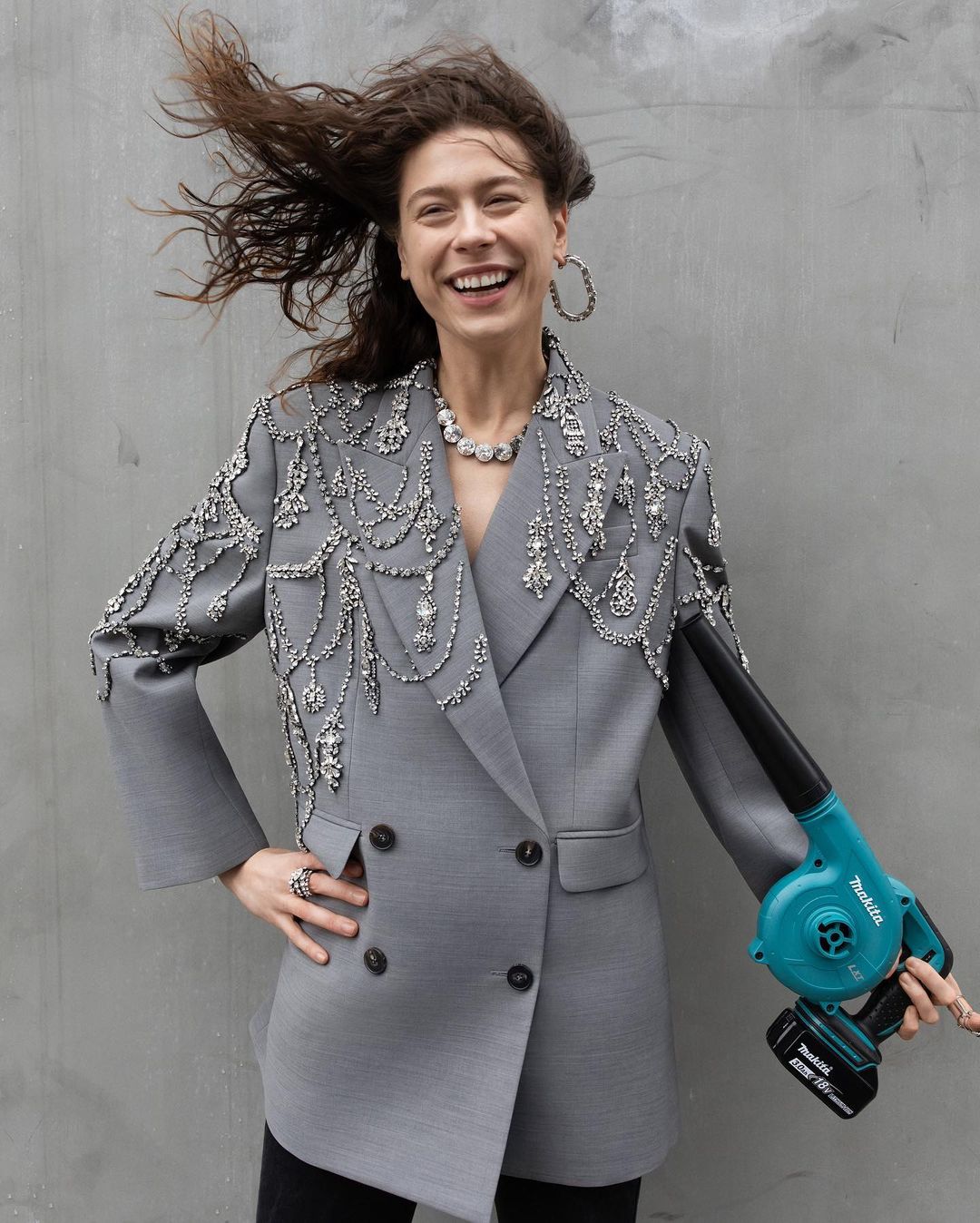 Woman in embellished grey blazer smiles, posing with a turquoise leaf blower against a plain grey background, creating a fashion editorial shot.