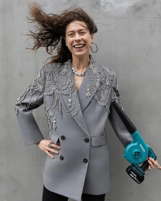Woman in embellished grey blazer smiles, posing with a turquoise leaf blower against a plain grey background, creating a fashion editorial shot.
