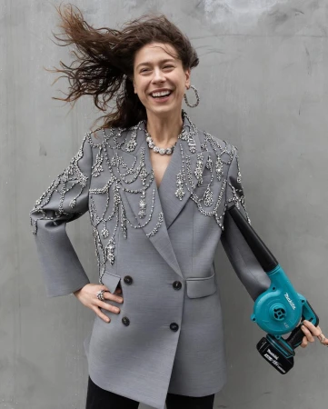 Woman in embellished grey blazer smiles, posing with a turquoise leaf blower against a plain grey background, creating a fashion editorial shot.