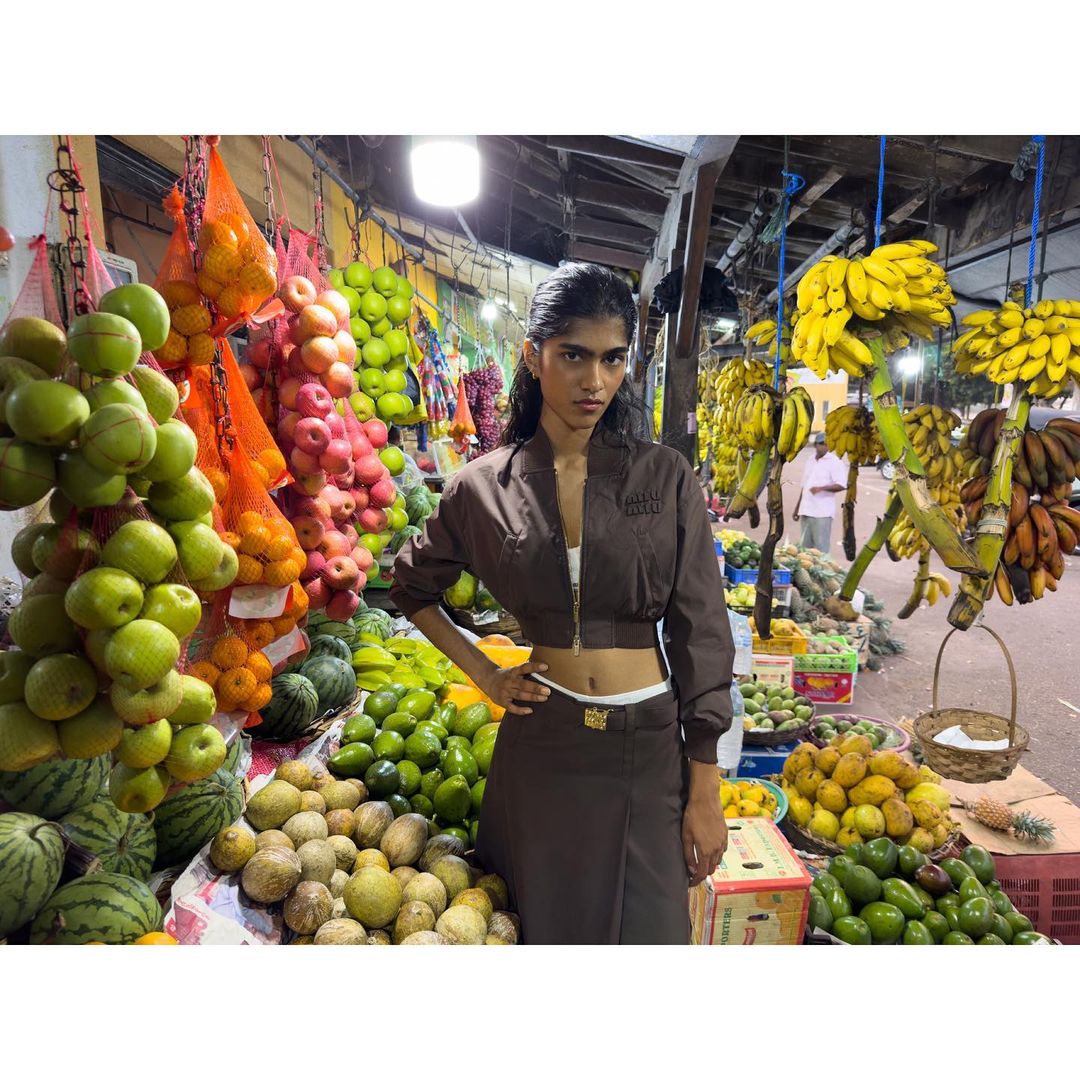 A young woman in a brown two-piece outfit poses in an outdoor market surrounded by colorful fruits, creating a fashion editorial.