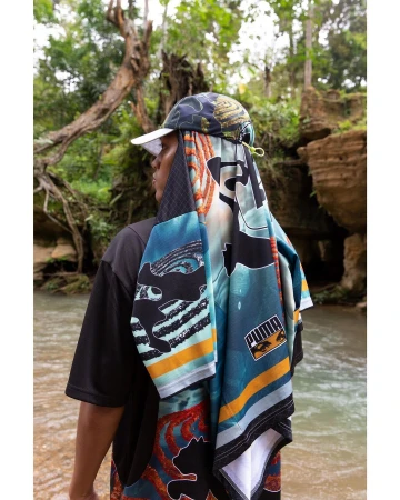 Person in nature setting wearing Puma-branded hat and patterned shawl, standing near a shallow stream surrounded by lush greenery.