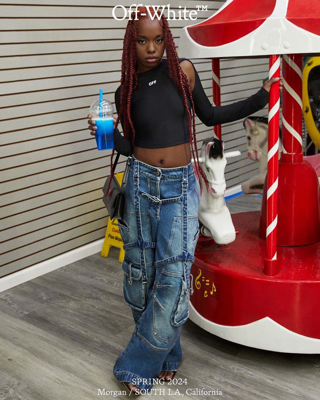 Young woman wearing Off-White, posing near a carousel with a blue slushie. She's wearing baggy jeans, a black crop top, and red braids.
