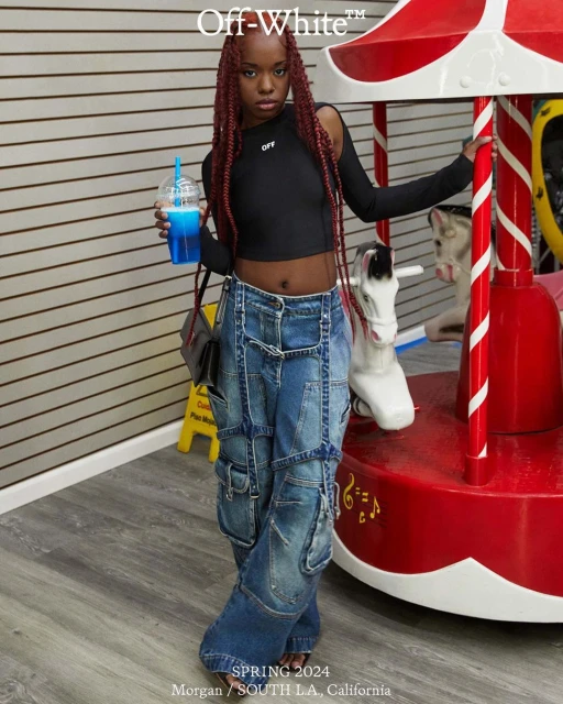 Young woman wearing Off-White, posing near a carousel with a blue slushie. She's wearing baggy jeans, a black crop top, and red braids.