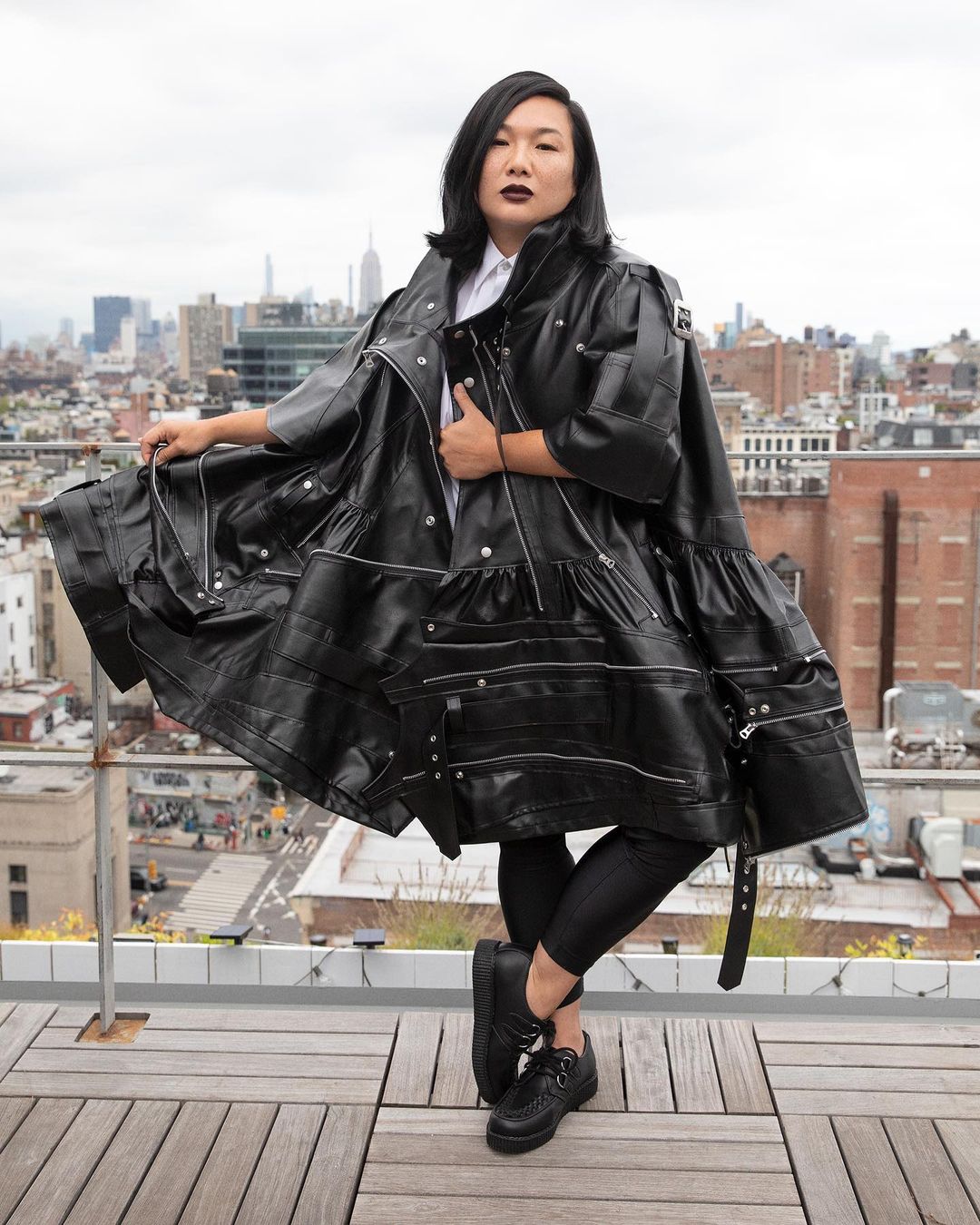 An Asian woman in an oversized black leather coat poses on a rooftop with a city skyline in the background on an overcast day.