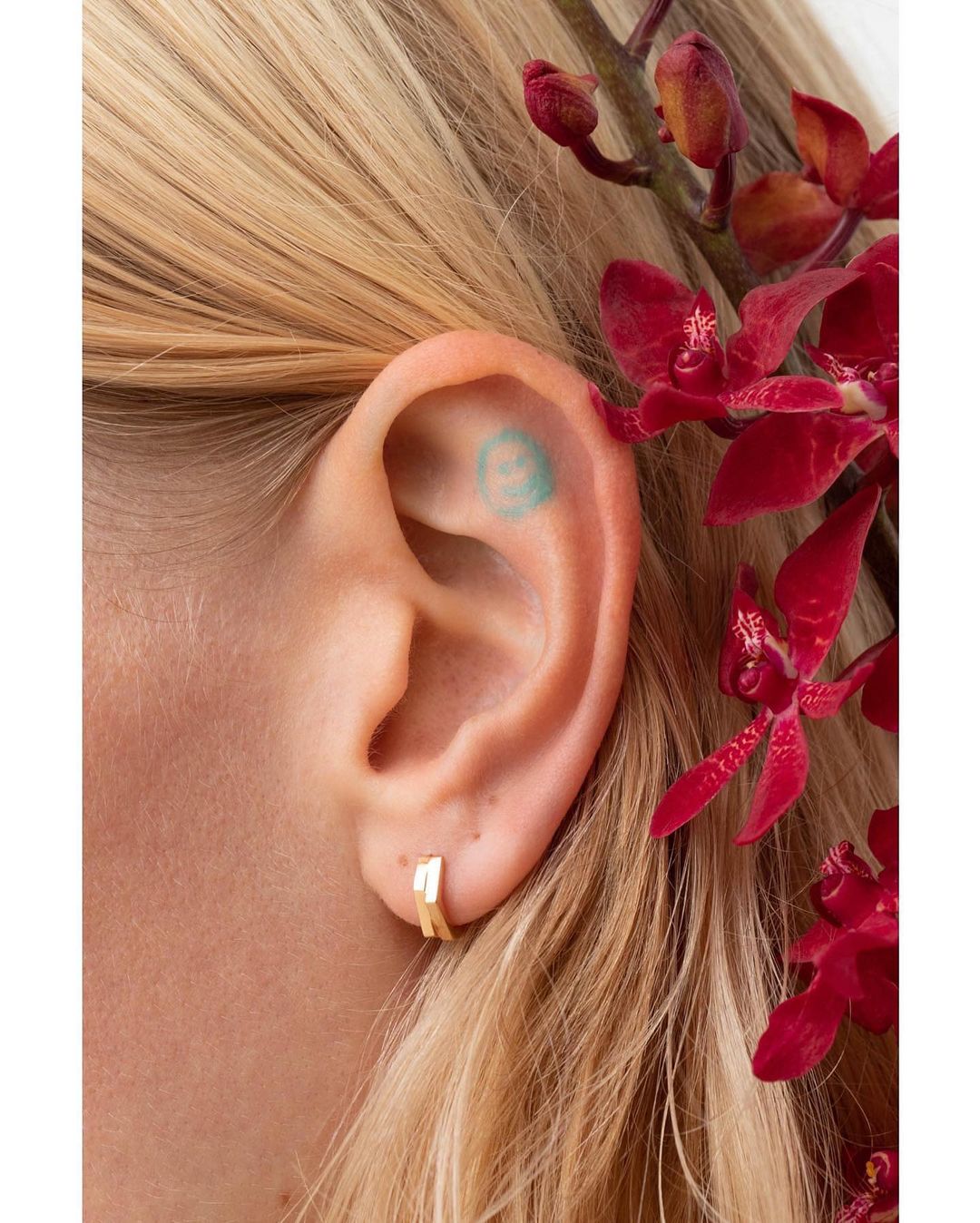 Close-up shot of a woman's ear adorned with a small gold hoop earring, a tiny tattoo inside the ear, and red flowers in the background.