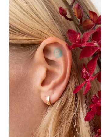 Close-up shot of a woman's ear adorned with a small gold hoop earring, a tiny tattoo inside the ear, and red flowers in the background.