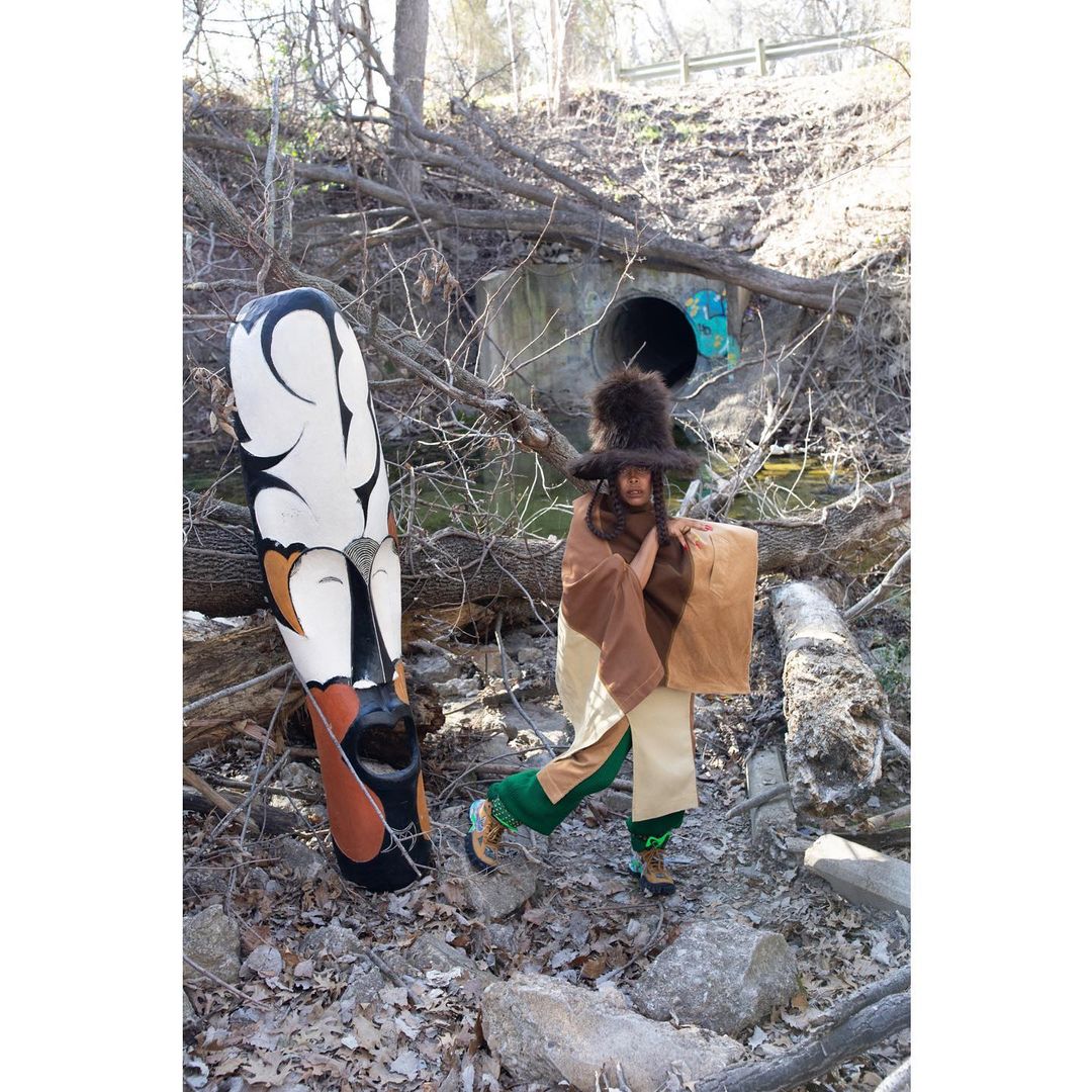 A person in costume stands next to a decorated pillar in a natural environment with dead tree branches and a stream flowing through a culvert.