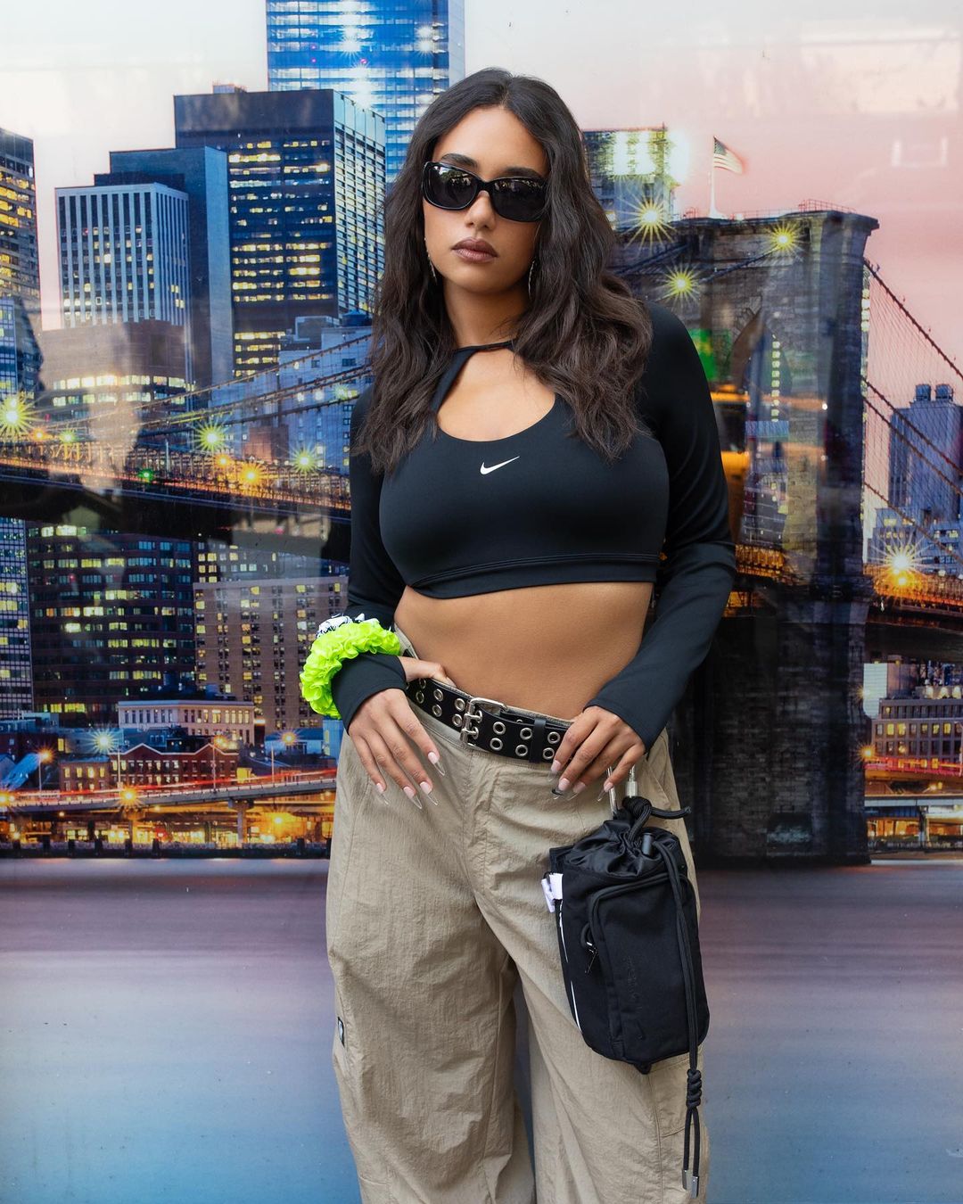 A woman with long dark hair wears a long-sleeve top, baggy pants, and sunglasses, posing in front of a cityscape backdrop.
