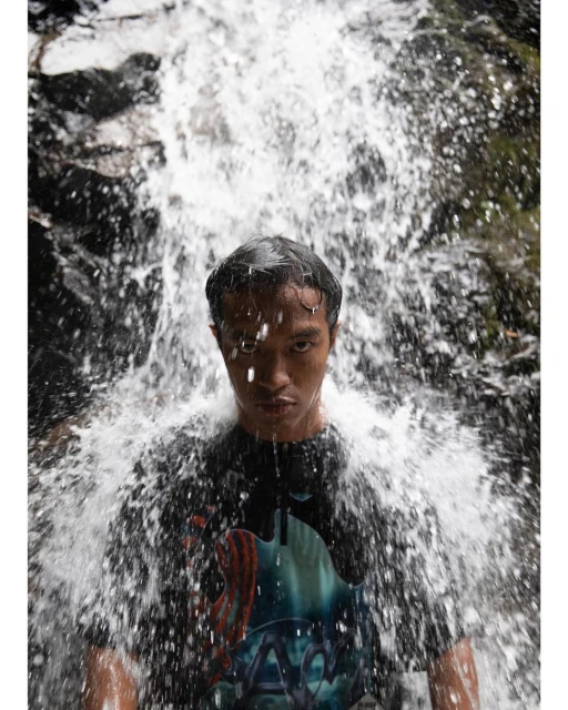 Portrait of a man in front of a waterfall with dark hair and a graphic t-shirt, looking directly at the camera with a serious expression.