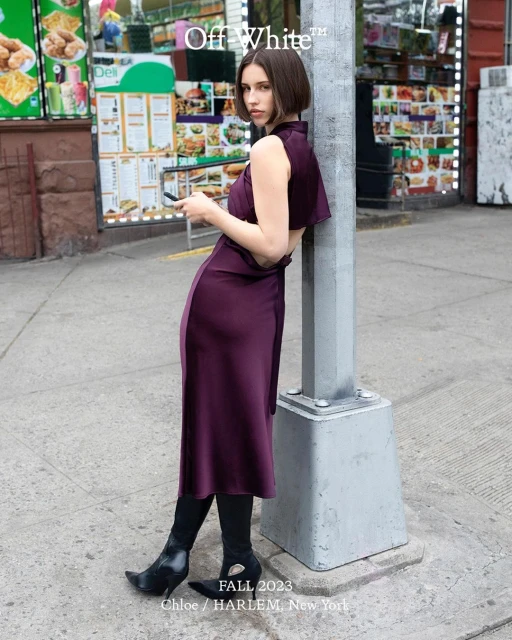 Woman in a stylish plum dress and black boots poses on a city sidewalk next to a streetlight, holding a phone, fall fashion.