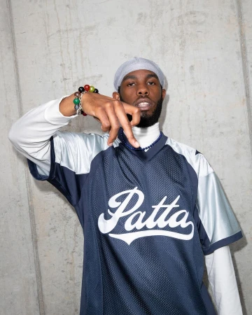 Portrait of a young Black man in a blue vintage-style jersey with the word 'Patta' against a gray concrete wall, wearing a gray head covering.
