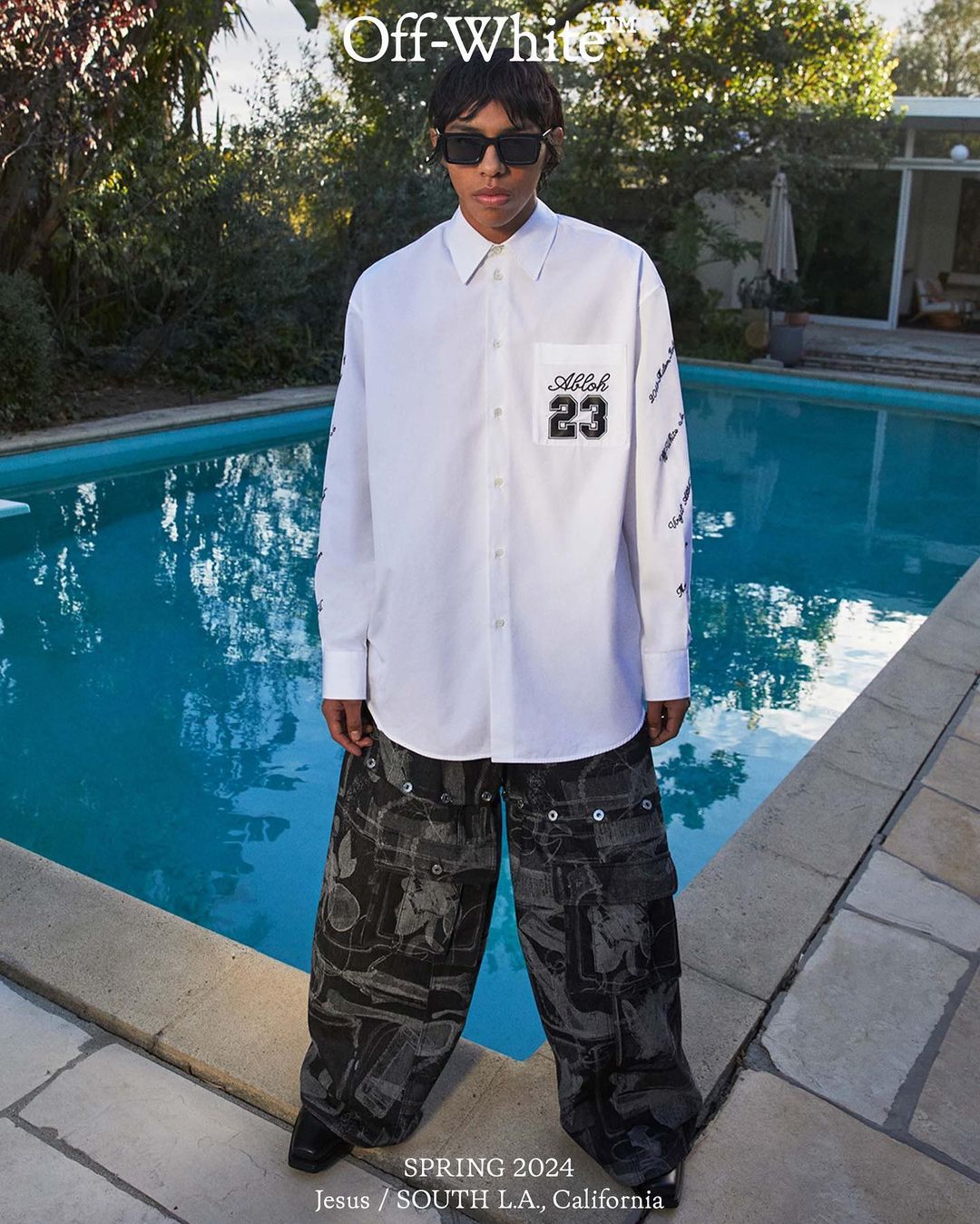 A person in a white long-sleeved shirt and patterned wide-leg pants stands by a turquoise pool outdoors with green trees behind them.