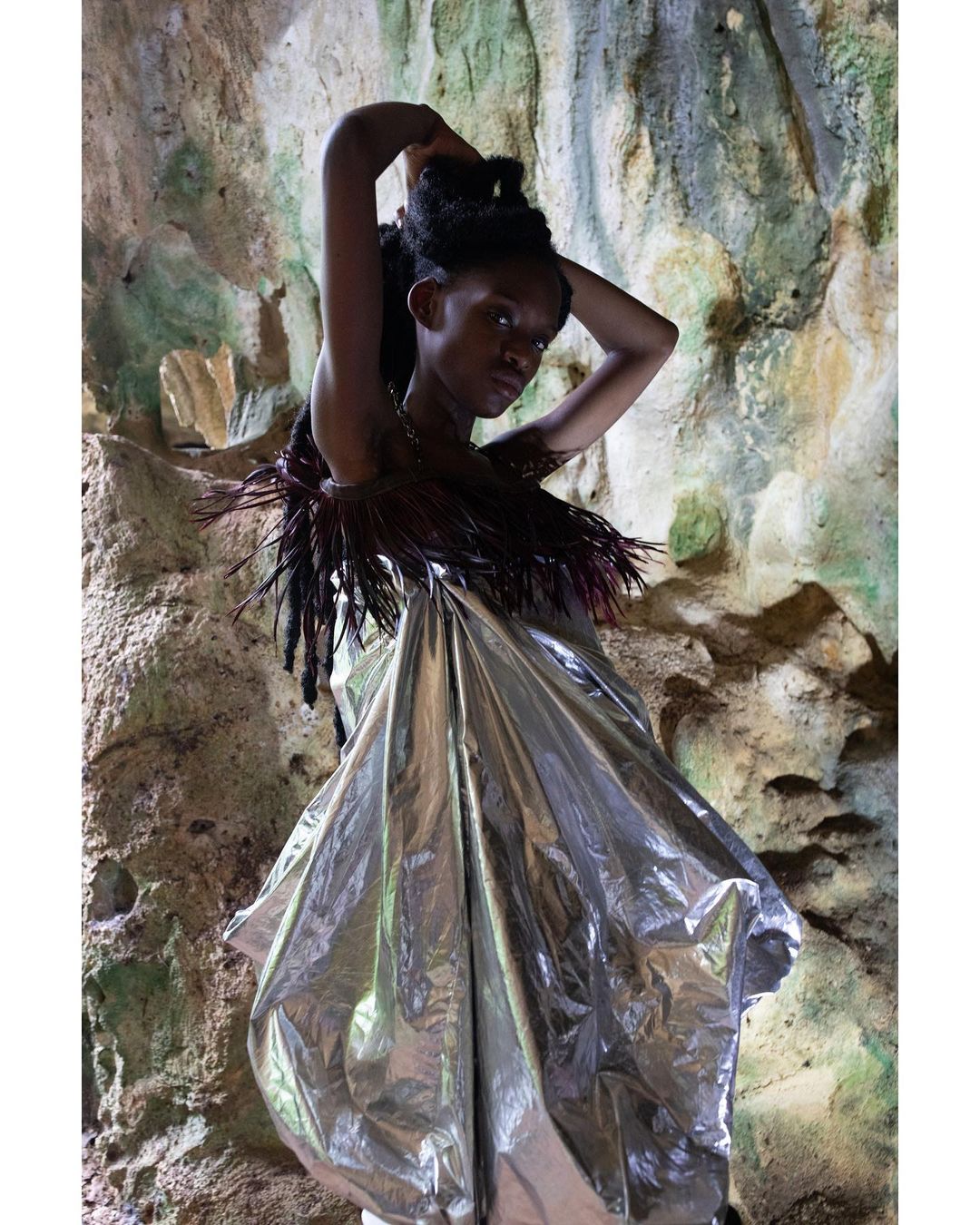 A striking portrait of a Black model in a metallic silver dress with feathered details posing inside a cave with rocky textures.