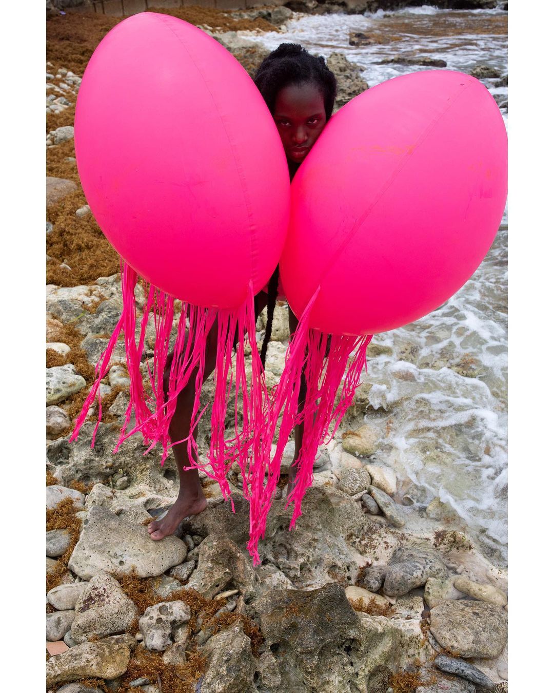 Woman stands on a rocky beach holding two large, bright pink, jellyfish-shaped balloons with trailing fringe on the ocean shore.