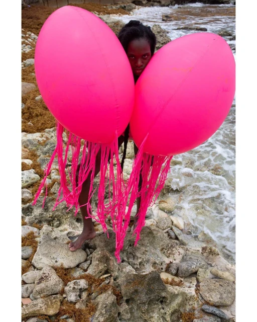 Woman stands on a rocky beach holding two large, bright pink, jellyfish-shaped balloons with trailing fringe on the ocean shore.