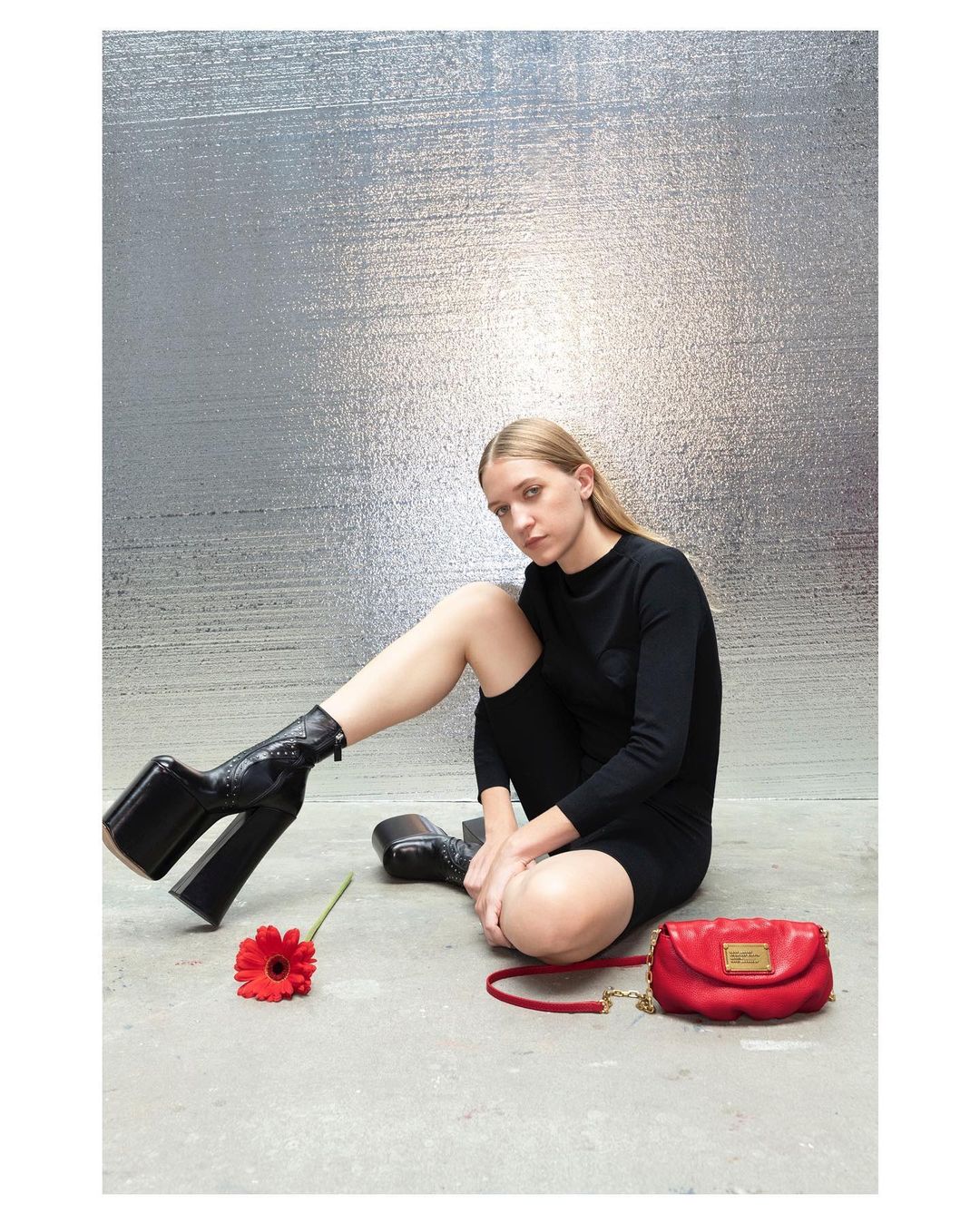 Fashion photography: Woman in black dress and platform boots sits next to a red purse and flower against a textured silver background.