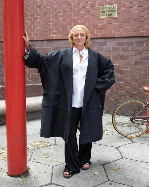 Adult female poses outdoors wearing an oversized black coat, white blouse and black pants in front of a red pillar and brick wall.