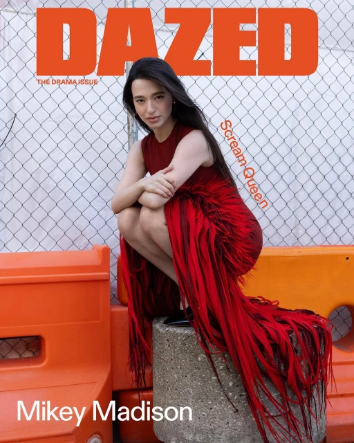 Mikey Madison poses for Dazed magazine cover in red dress. Woman crouches on cement block in front of orange barriers, chain-link fence backdrop.