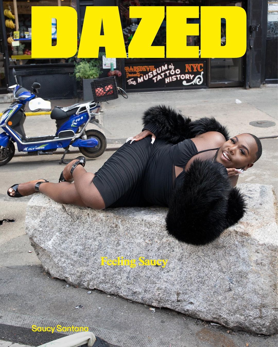 Magazine cover of Dazed featuring an adult posing on a stone, wearing a black dress and black faux fur shawl, with a blue scooter.
