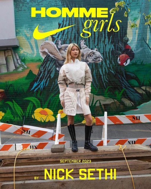 Editorial fashion shot of a woman in streetwear posing in front of a mural with construction barriers, featuring Nike branding and a street style aesthetic.